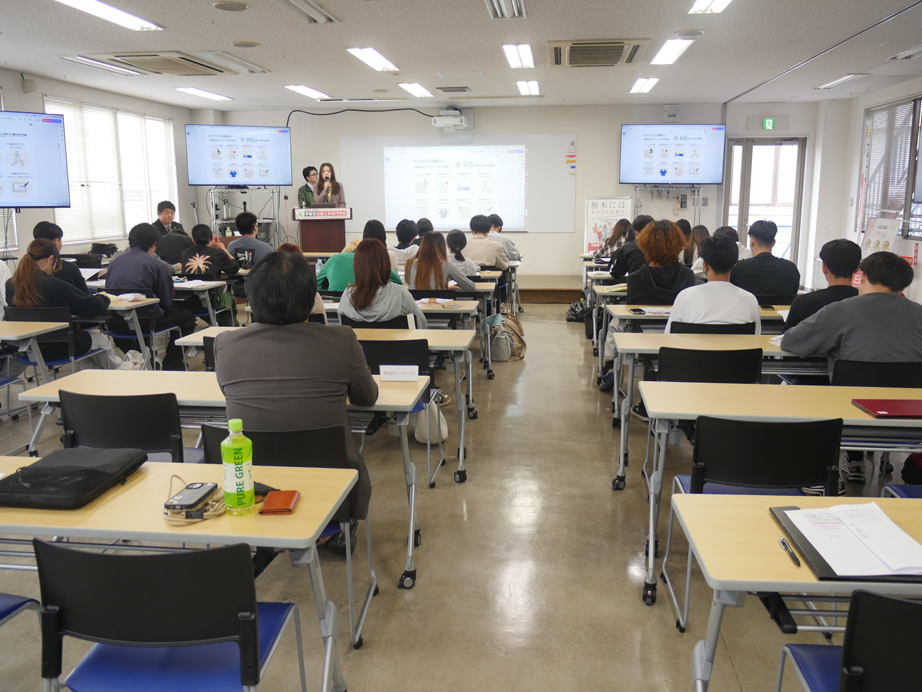 企業担当者と卒業生が登壇し、会社概要をスクリーンに映しながら説明している様子